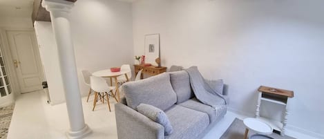 Uma sala de estar com um sofá cinzento, uma mesa redonda pequena e uma cadeira branca.