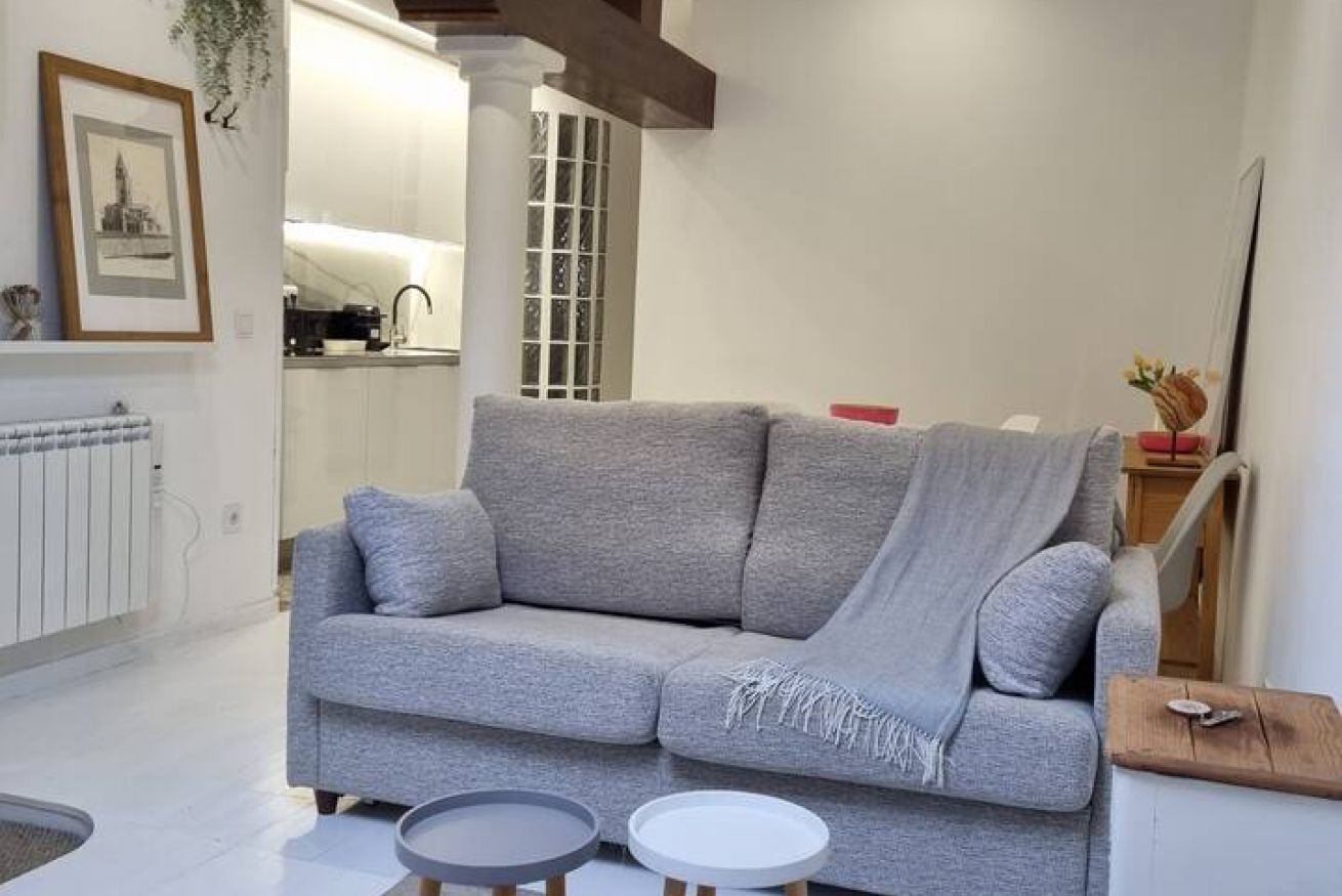 Ein Wohnzimmer mit grauem Sofa, zwei runden Beistelltischen, einer Heizung, einem gerahmten Bild und einer Küche im Hintergrund.