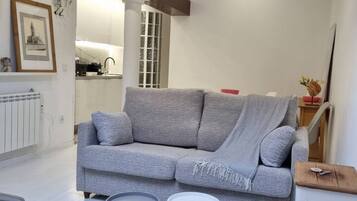 Ein Wohnzimmer mit grauem Sofa, zwei runden Beistelltischen, einer Heizung, einem gerahmten Bild und einer Küche im Hintergrund.