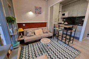 A compact living space with a sofa, a small round table, a kitchen area with cabinets and a microwave, and a patterned rug.