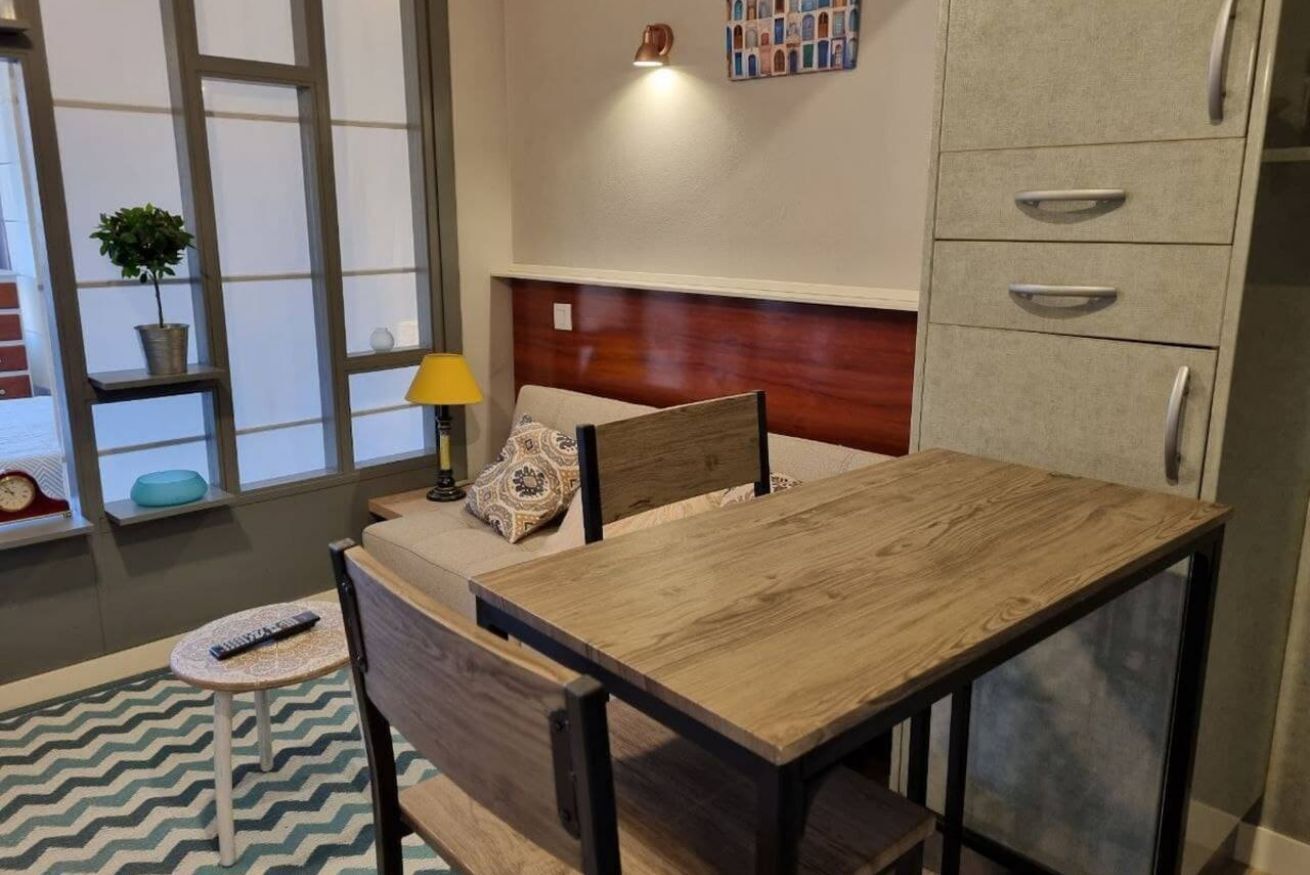 A small, modern room with a wooden table, a bed with a headboard, a lamp, a potted plant, and a wall-mounted shelf.