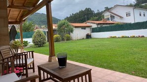 Um pátio ao ar livre com mesa e cadeiras, um copo de bebida e vista para um gramado verde e edifícios ao fundo.