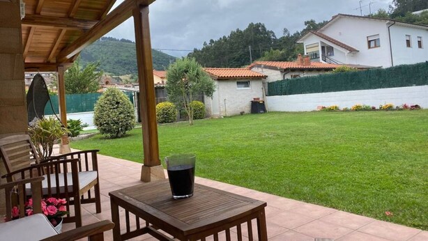 Une terrasse extérieure avec une table et des chaises, un verre rempli d’une boisson, et une vue sur une pelouse verdoyante et des bâtiments en arrière-plan.