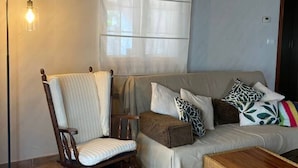 Un salon avec un fauteuil en bois, un canapé agrémenté de coussins à motifs, une lampe sur pied et une fenêtre ornée de rideaux légers.