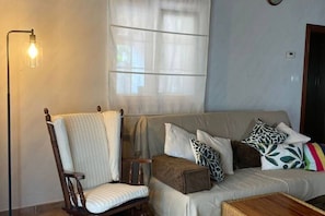 A living room with a wooden chair, a sofa with patterned cushions, a floor lamp, and a window with sheer curtains.