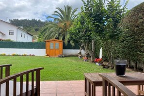 Eine Terrasse mit Holztisch und einem Glas Wasser, eine kleine Holzscheune und ein gepflegter Rasen mit verschiedenen Pflanzen und Blumen.
