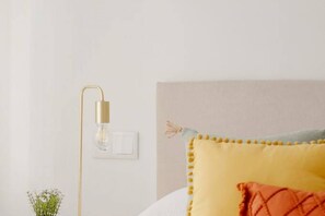 A bed with a yellow headboard and a single orange pillow, a bedside lamp, and a small plant on the nightstand.