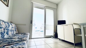A small, sparsely furnished room with a sofa, a television, and a sliding glass door leading to an outdoor area. - Modern Apartment Near the Beach in Jesolo, Italy (Jesolo)