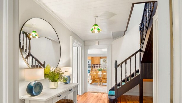 A hallway with a wooden floor, a white console table with a mirror, a lamp, and a potted plant. There is a staircase with a dark railing leading to another room. - Sycamore Cottage on the outskirts of Clifden (Clifden)