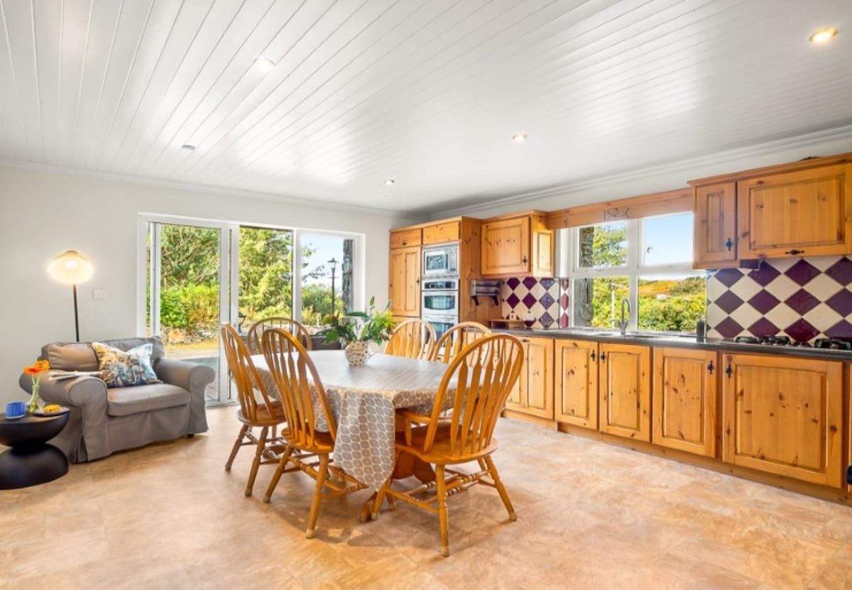 A modern kitchen with wooden cabinets, a dining table with chairs, and a view of greenery outside.