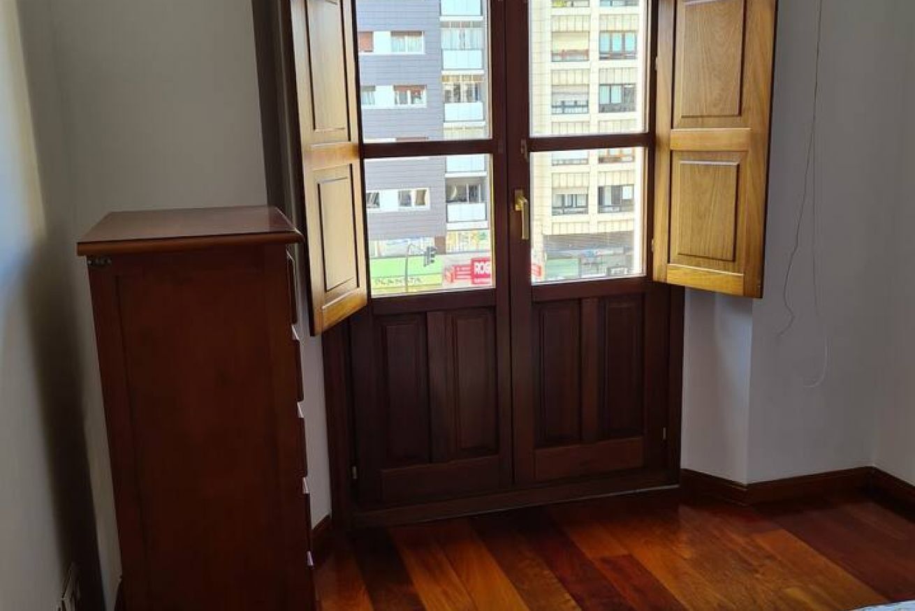 Habitación de esquina con un armario de madera, una puerta de madera con paneles de vidrio y vista a un edificio a través de la puerta.