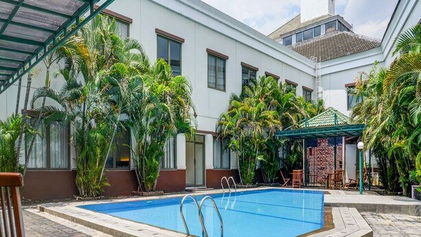 A swimming pool with a glass roof, surrounded by palm trees and a white building with multiple windows. - Townhouse OAK Hotel Grand Cikarang (Cikarang)