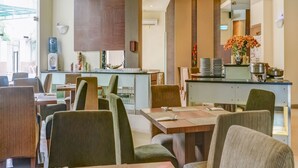 A modern dining area with high tables and comfortable chairs, a water dispenser, and a kitchen counter with utensils and plates. - Townhouse OAK Hotel Grand Cikarang (Cikarang)