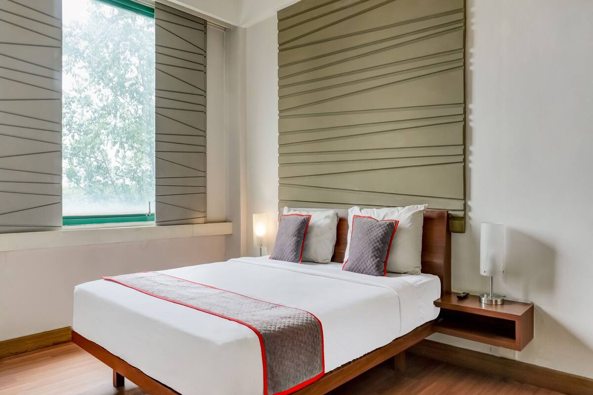 A modern hotel room with a large bed, a bedside table, a nightstand, and a window with blinds.