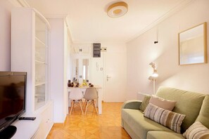 Ein Wohnzimmer mit Fernseher, einem grünen Sofa, einem Esstisch mit Stühlen und einer Küchenzeile inklusive Schränken und Kühlschrank.