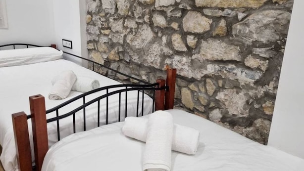A room with two beds, each with white linens and wooden headboards. A stone wall is visible behind the beds.