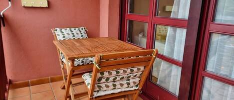 A wooden table and chairs with patterned cushions, a red-framed window, and a tiled floor.