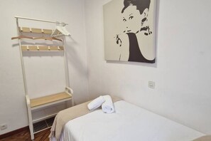 A single bed with a white mattress and a rolled white towel, a white wall-mounted radiator, and a white metal bunk bed with wooden slats.