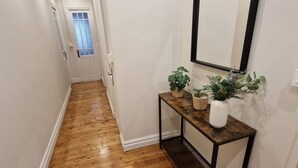 Un couloir doté d’un plancher en bois, une porte blanche au fond, et une console ornée de plantes et d’un vase.
