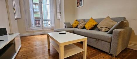 A living room with a grey sofa, a white coffee table, wooden flooring, and a balcony with open doors.