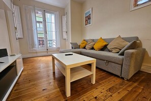 Una sala de estar con un sofá gris, una mesa de centro blanca, piso de madera y un balcón con puertas abiertas.