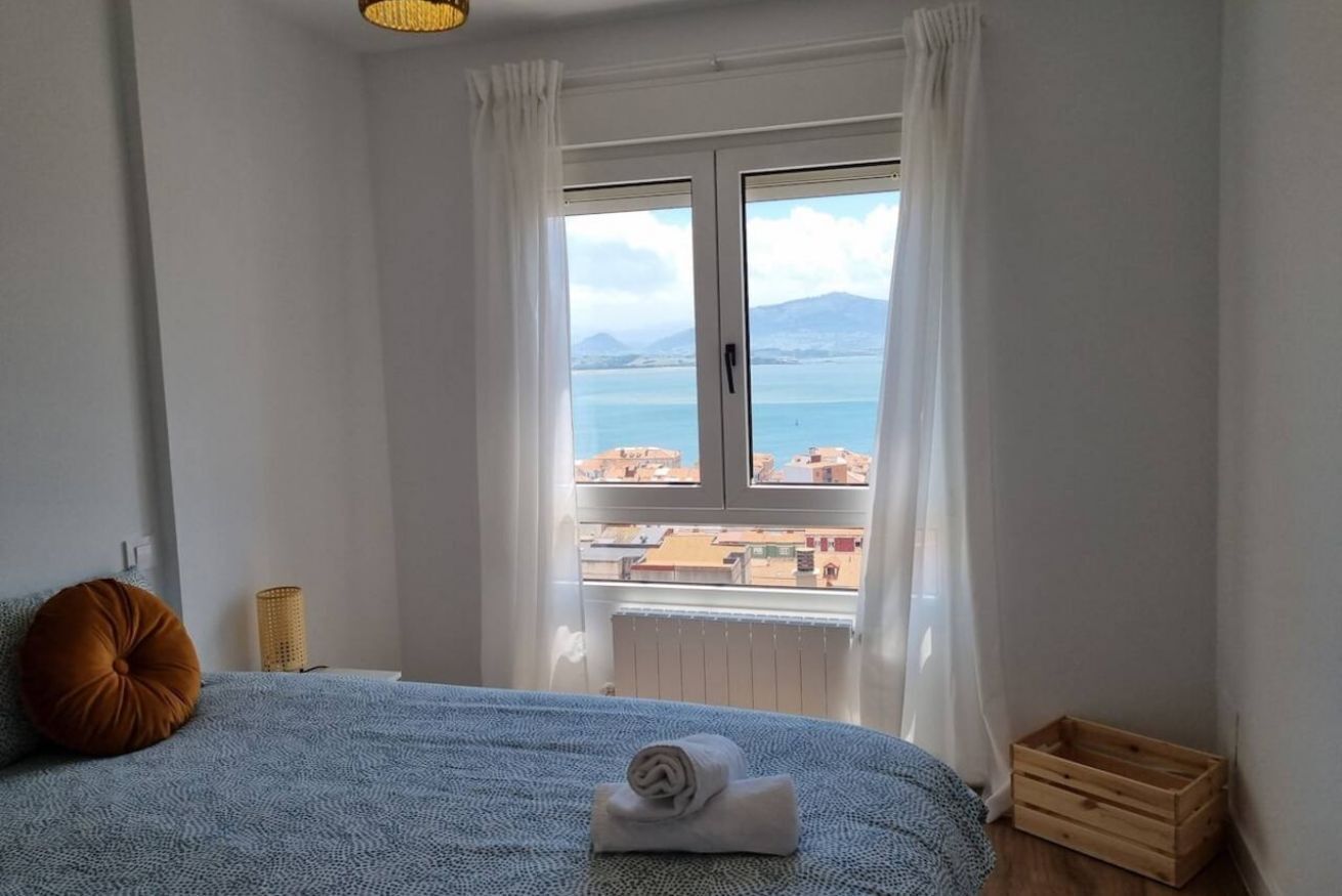 Ein Schlafzimmer mit einem Bett, einem Fenster mit Vorhängen, Blick auf das Meer und einer Holzkiste.
