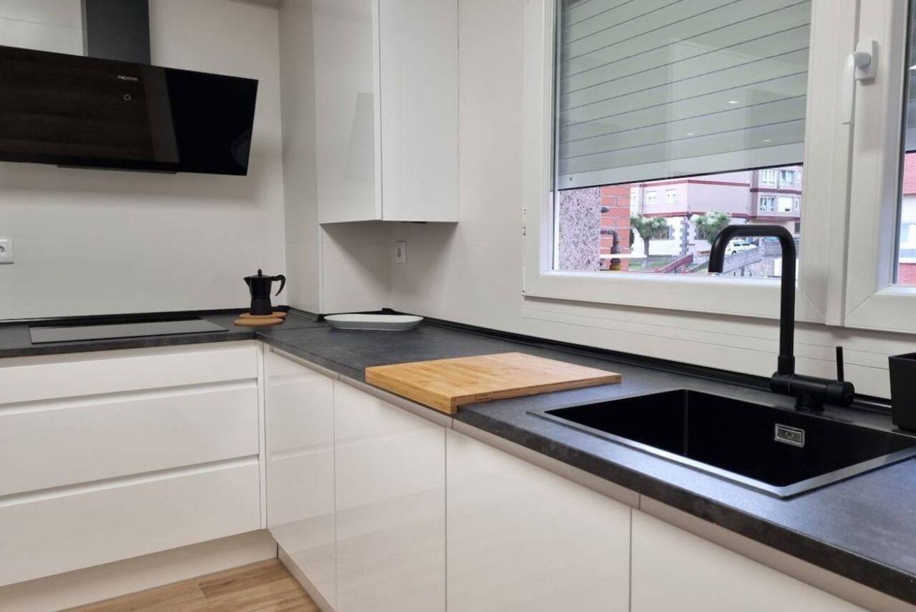 A modern kitchen with white cabinets, a black countertop, a wooden cutting board, a black sink, and a window with a view of buildings.