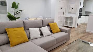 A modern living room with a grey sofa, a glass coffee table, and a dining area in the background.