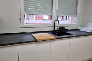 Une cuisine avec un plan de travail noir, une planche à découper en bois, un évier et une fenêtre avec des stores.