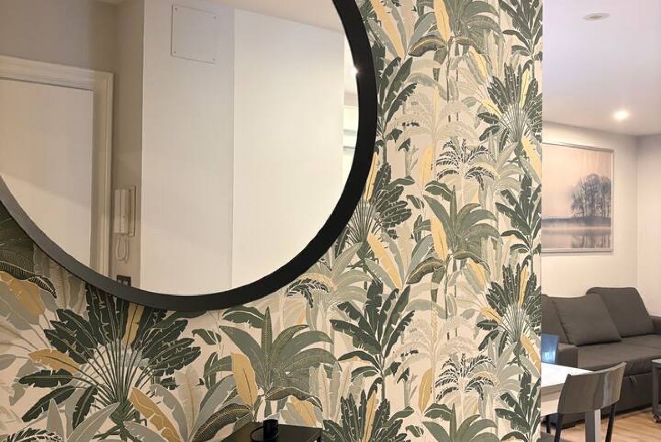 A room with a round mirror, a patterned wallpaper, a grey sofa, and a table.