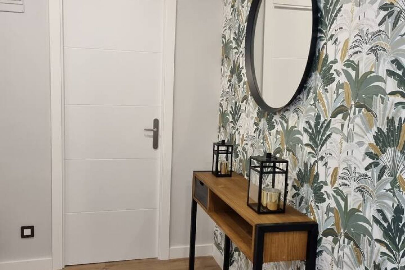 A hallway with a white door, a wooden console table with two black lanterns, and a wall with a floral wallpaper pattern.