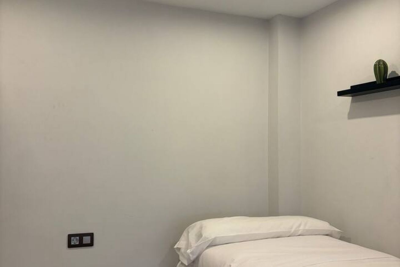 A small, single-bed room with a white wall, a black shelf with a cactus, and a power outlet.