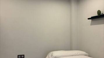 A small, single-bed room with a white wall, a black shelf with a cactus, and a power outlet.