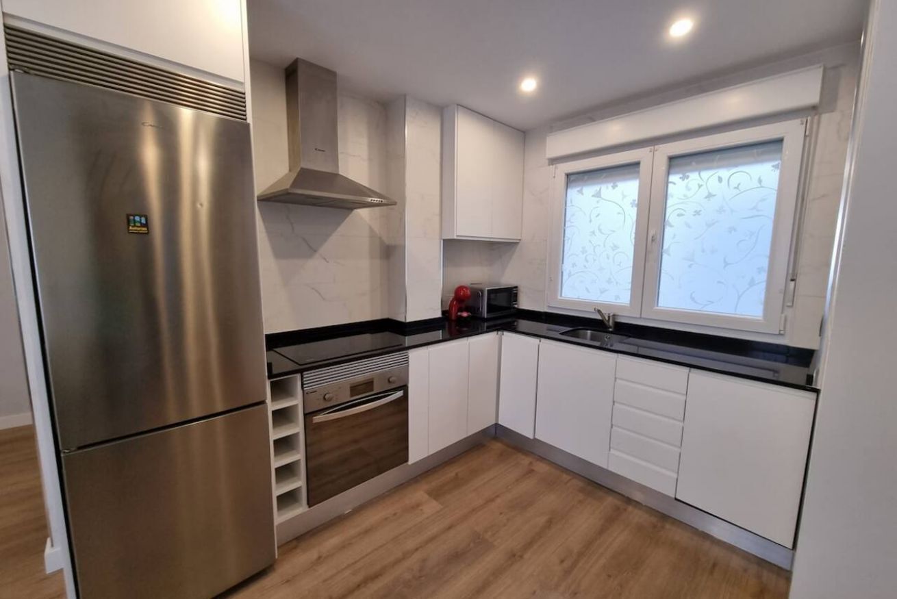 A modern kitchen with white cabinetry, a stainless steel refrigerator, a built-in oven, and a window with frosted glass.