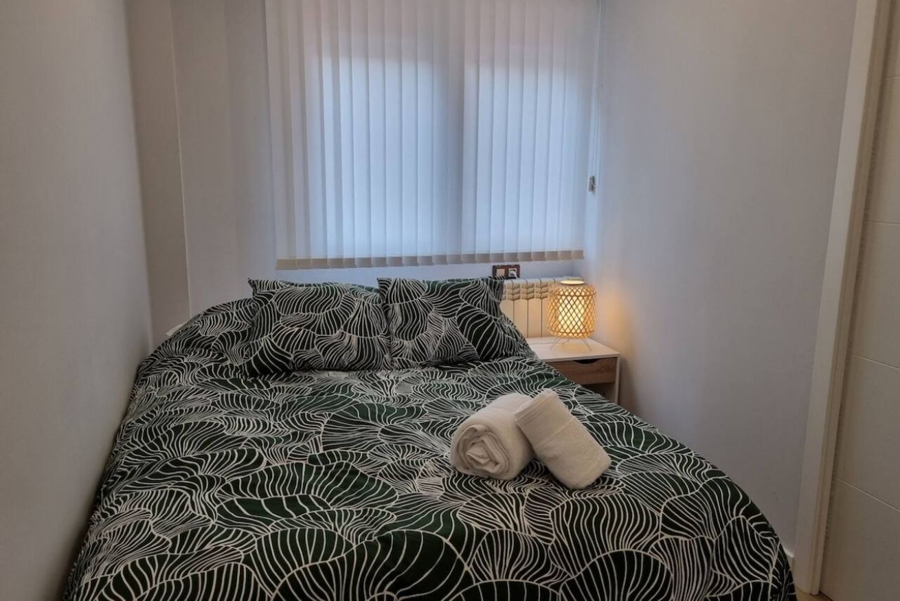 Una cama con una colcha estampada, una toalla enrollada, una lámpara de noche y una ventana con persianas.