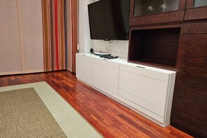 Une pièce dotée d’un plancher en bois, d’un meuble de rangement blanc abritant un téléviseur à écran plat, et d’une grande fenêtre avec des rideaux.