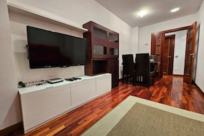 A modern living room with a flat-screen TV mounted on a white cabinet, a wooden cabinet with glass doors, a dining area with a black table and chairs, and a wooden floor. - 604a - Modern 2-Bedroom Apartment by R2R (Oviedo)