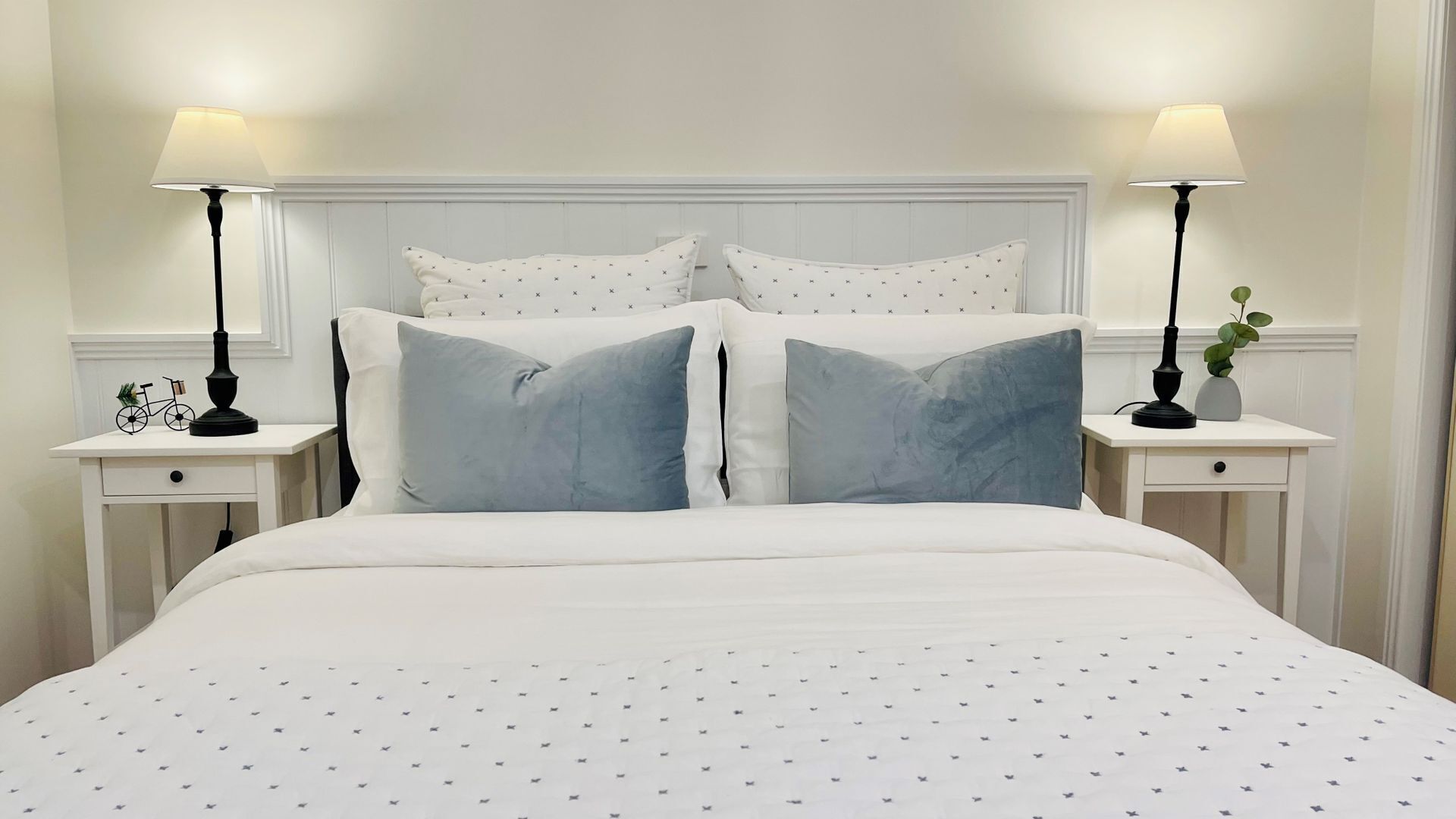 Cama bem arrumada com edredom branco e almofadas azuis, acompanhada por duas mesas de cabeceira com abajures e uma pequena planta.