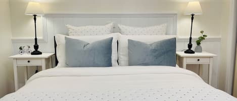 Cama bem arrumada com edredom branco e almofadas azuis, acompanhada por duas mesas de cabeceira com abajures e uma pequena planta.