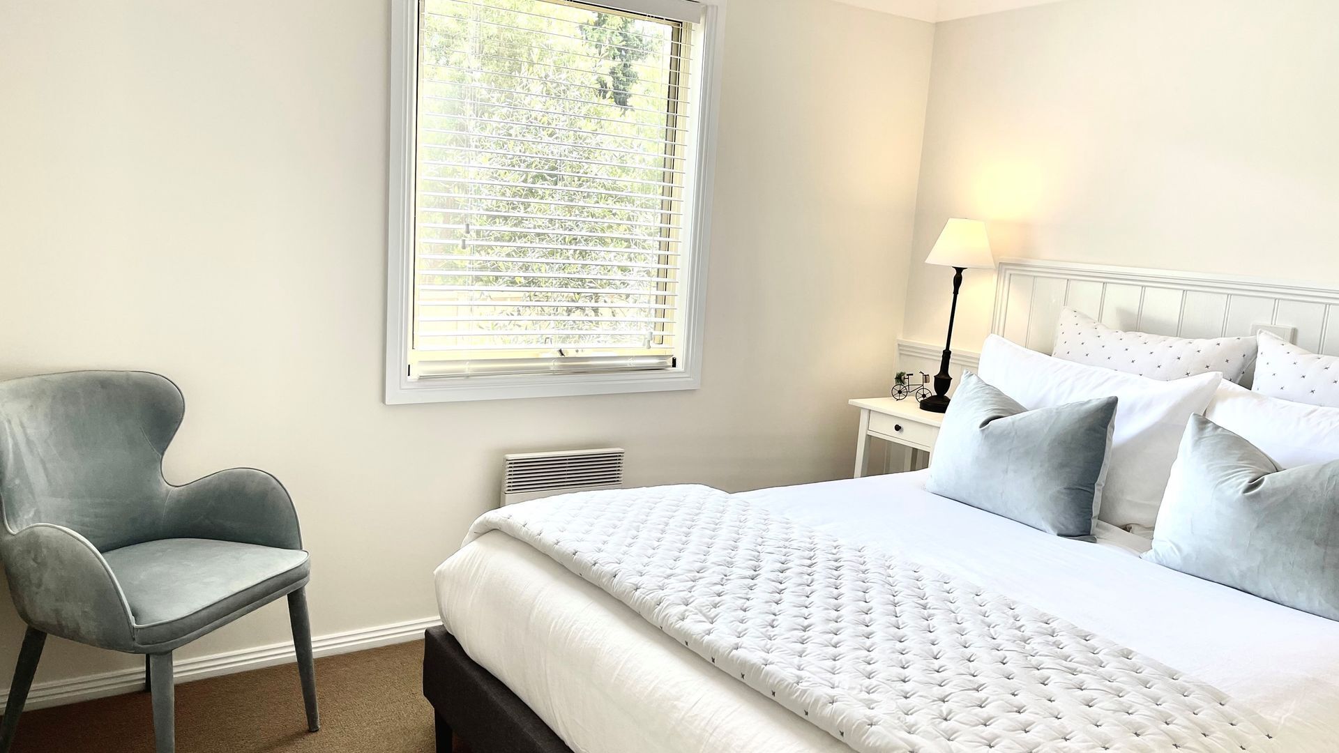 A hotel room with a bed, a chair, a nightstand, a lamp, and a window with blinds.