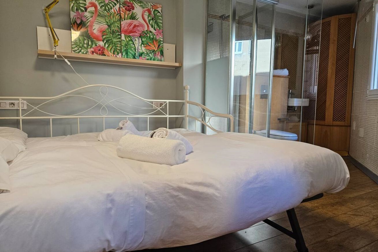 A neatly made bed with white linens and towels, a wall-mounted shelf with a painting, and a glass shower enclosure.