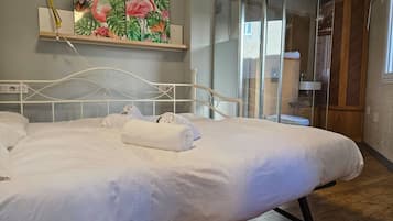 Una cama bien hecha con sábanas y toallas blancas, un estante en la pared con un cuadro y un box de ducha de vidrio.