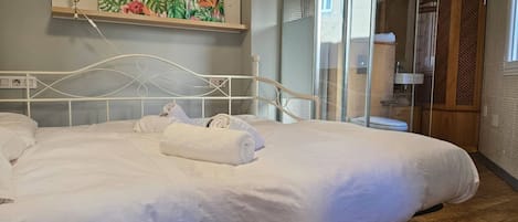 A neatly made bed with white linens and towels, a wall-mounted shelf with a painting, and a glass shower enclosure.