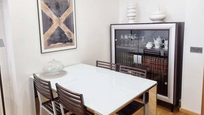 Un comedor con una mesa blanca, sillas de madera, un centro de mesa de vidrio, un cuadro abstracto enmarco y un mueble con puertas de cristal que contiene utensilios de cocina.