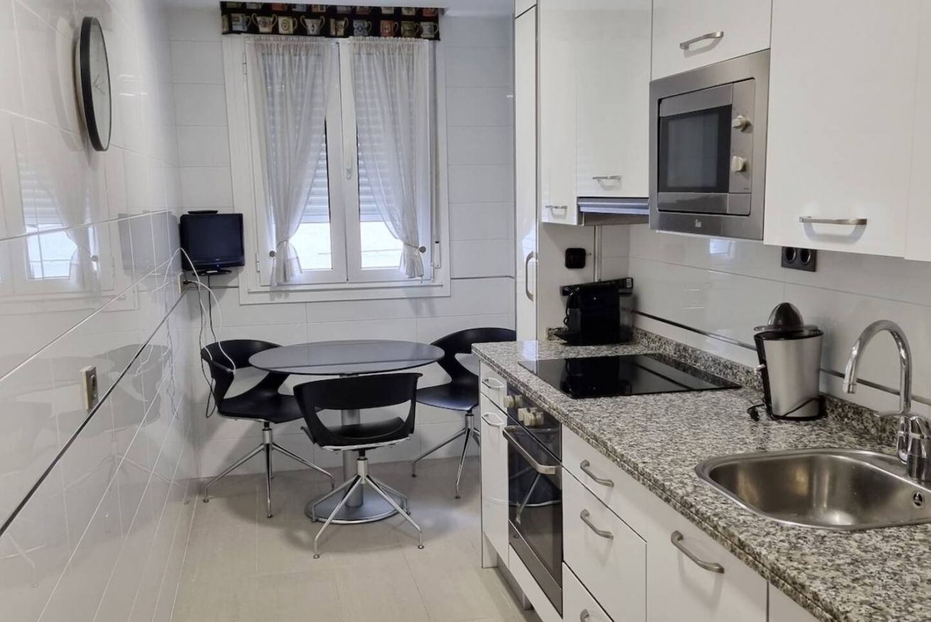 Une cuisine moderne avec des meubles blancs, un plan de travail en granit, un four encastré, une machine à café et une fenêtre avec des rideaux.