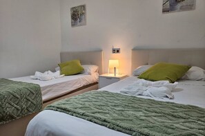 A hotel room with two beds, each with a green blanket, white linens, and a nightstand with a lamp.