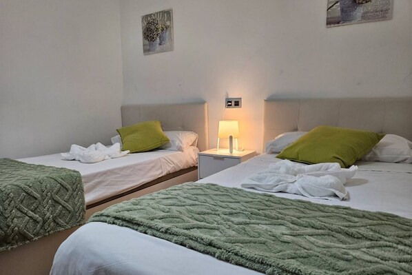 A hotel room with two beds, each with a green blanket, white linens, and a nightstand with a lamp.