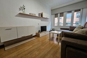 A modern living room with a sofa, TV, wooden flooring, and a window with a view of buildings.