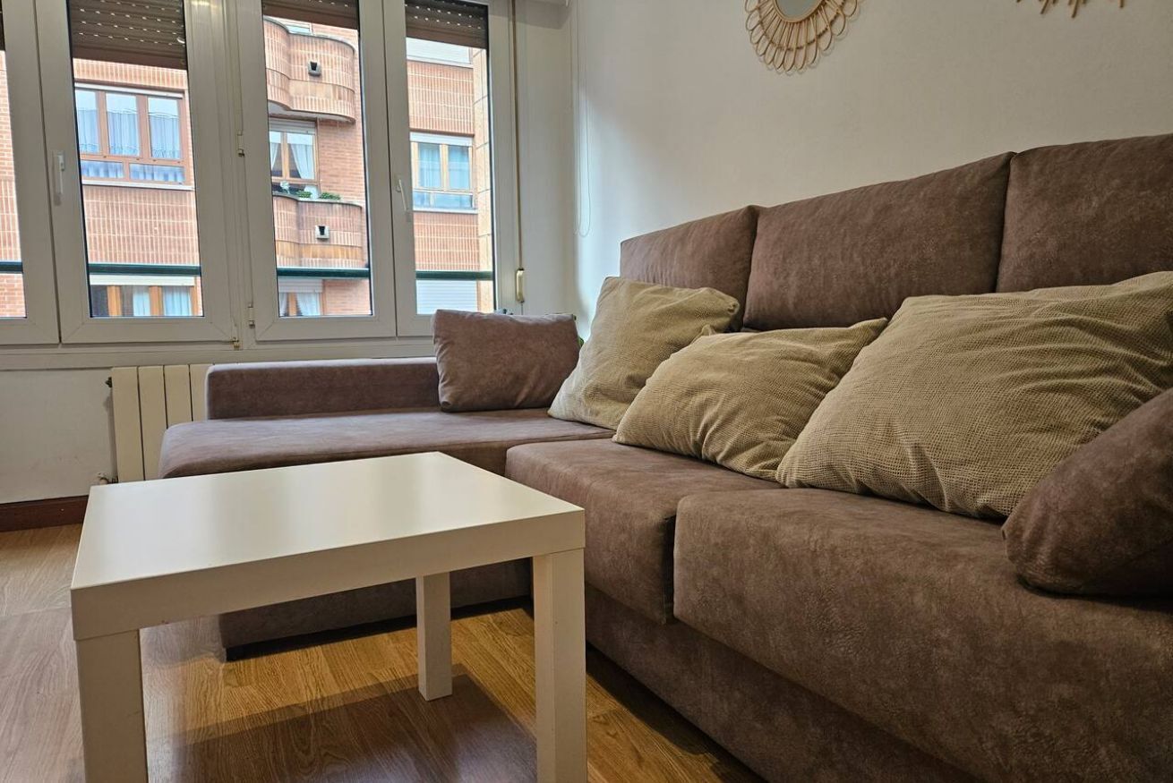 Un salon avec un canapé marron, une table blanche et une vue sur un bâtiment en briques, visible à travers une fenêtre.