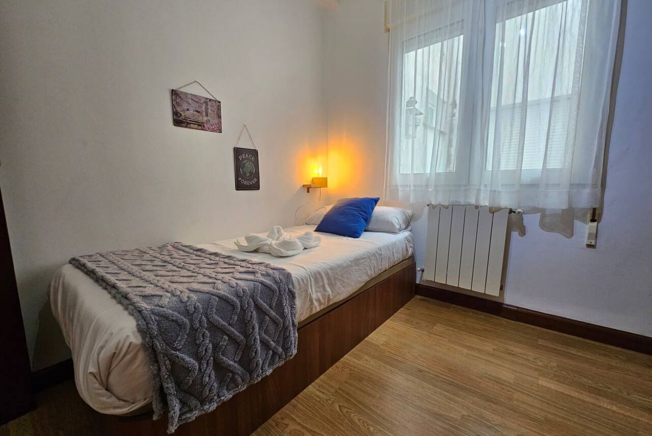 A single bed with a blanket, a bedside lamp, and a window with sheer curtains.